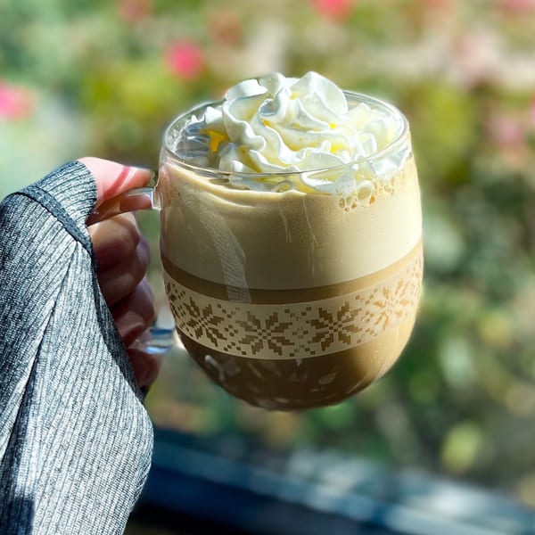 A hand in a gray sleeve holds a BarleyBean coffee mug with whipped cream, outdoors.