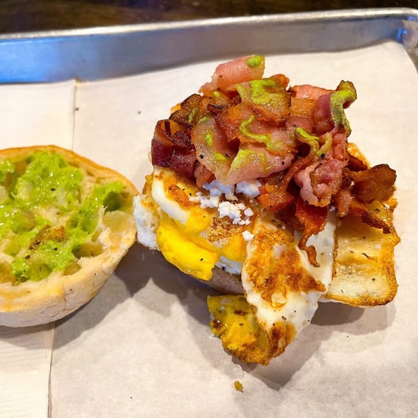 An open-faced breakfast sandwich with avocado spread, fried eggs, bacon, and feta cheese.
