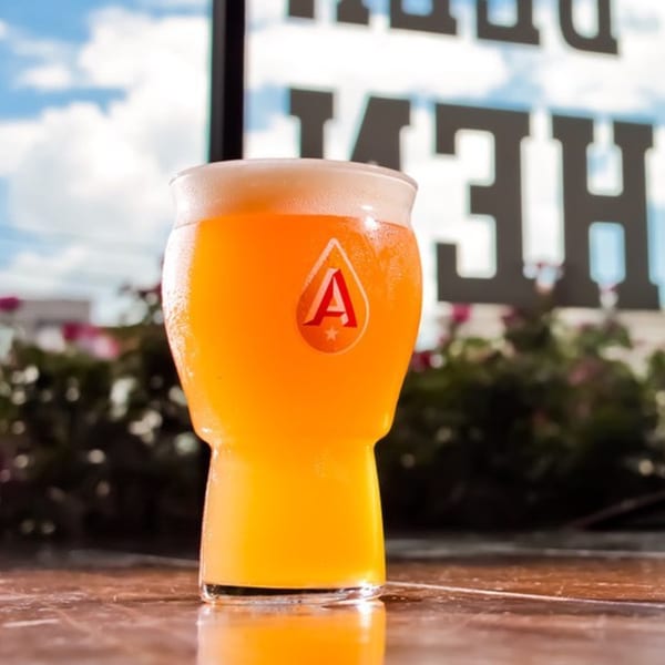 A glass of foamy orange beer with a red "A" logo sits on a table with a cloudy sky background.