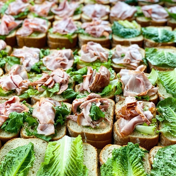 Rows of open-faced sandwiches with ham and lettuce on rustic bread, ready for catering at BarleyBean.