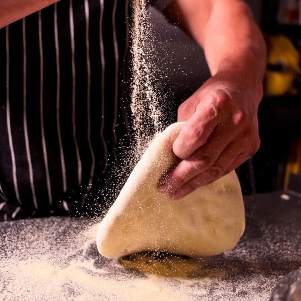 A chef's hands dusting flour onto a piece of dough on a dark surface.