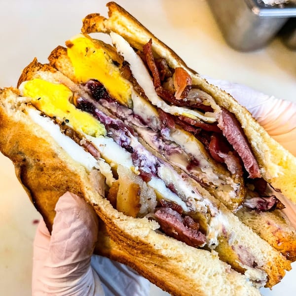 A hand in a white glove holds a hearty breakfast sandwich with egg, bacon, and jam.