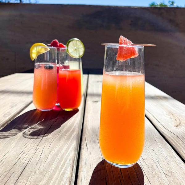 Three colorful BarleyBean cafe drinks with fruit garnishes sit on a sunlit wooden table.