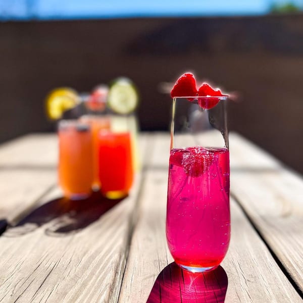 Bright pink mimosa with raspberries on a wooden table, with other colorful drinks blurred in the background.