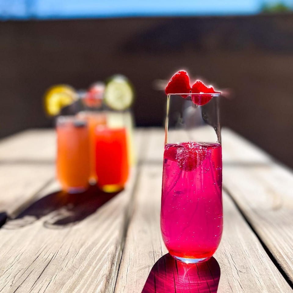 Bright pink mimosa with raspberries on a wooden table, with other colorful drinks blurred in the background.