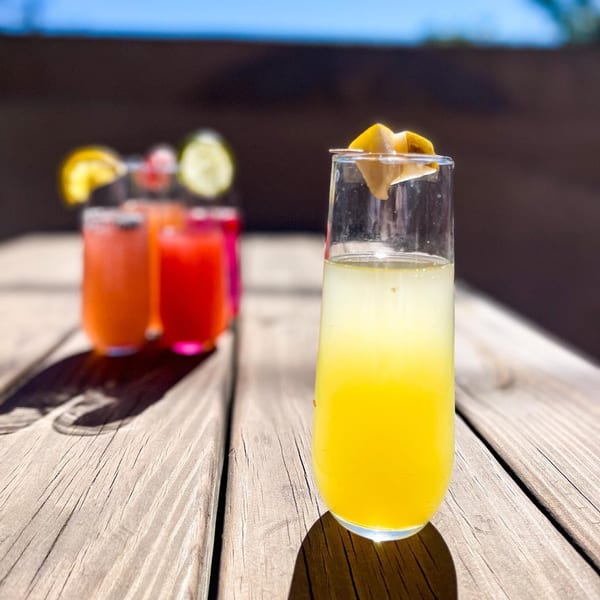 A bright yellow drink with a lemon garnish sits on a wooden table, with other colorful drinks blurred in the background.