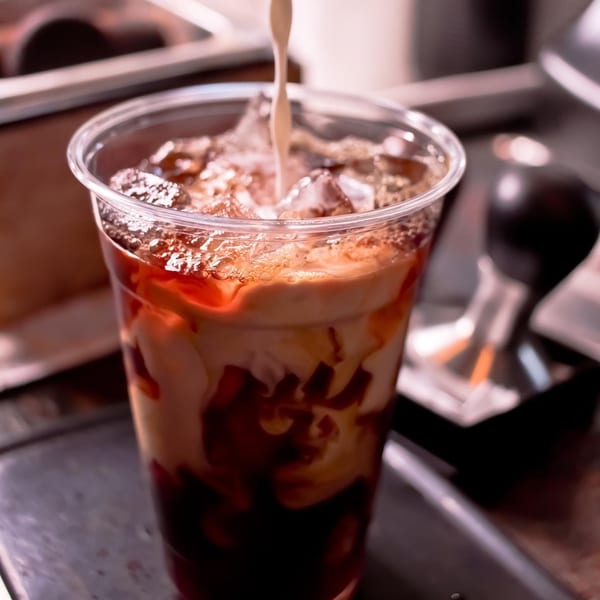 Milk being poured into an iced coffee with chocolate syrup in a clear plastic cup.