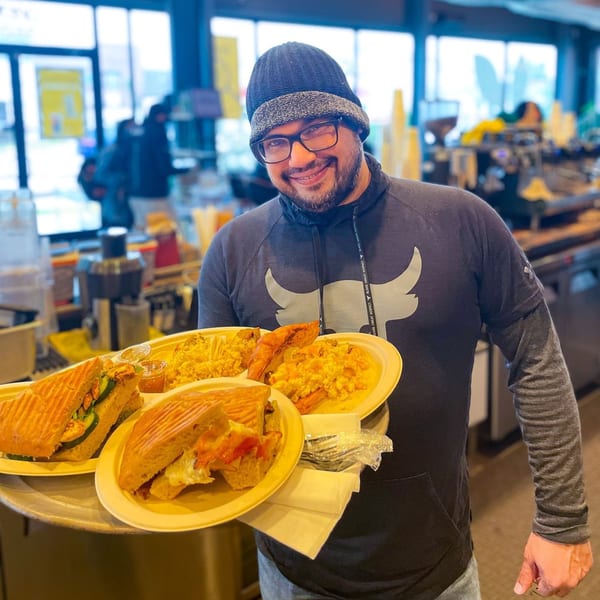 A smiling man in a beanie and glasses holds a tray with paninis and scrambled eggs at BarleyBean.