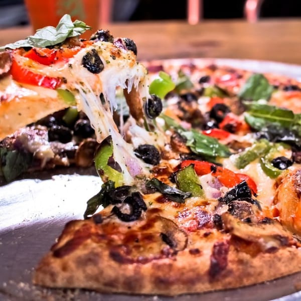 A slice of BarleyBean's veggie pizza with melted cheese being lifted from the whole pizza.